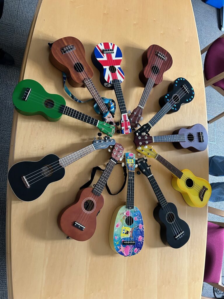 Collection of small colorful ukuleles laid on a wooden table, including a Union Jack design and a green body. | Inclusion School Aldershot Thank you to Romsey Ukulele Group | Inclusion School Aldershot Thank you to Romsey Ukulele Group